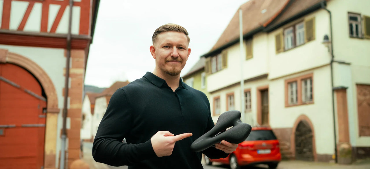 Mann in schwarzem Pullover zeigt in einer Fachwerk-Altstadt auf den Alpensattel in seiner Hand